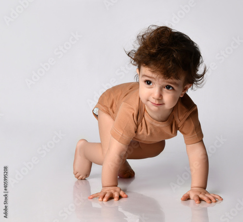 Φωτογραφία charming dark-haired curly-haired boy crawls on all fours on a gray background