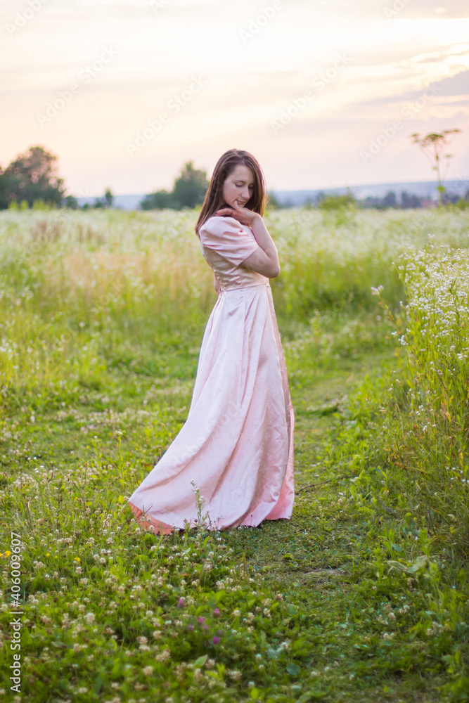 Beautiful photo of woman with dark hair standing in the wildflowers field barefoot holding the bottom of the long pink dress on a warm summer evening. Joyfull summer walking. Beautiful photo of woman with dark hair standing in the wildflowers field barefoot holding the bottom of the long pink dress on a warm summer evening. Joyfull summer walking.