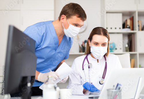 Two physicians wearing medical masks and protective gloves working with case histories on laptop in modern office