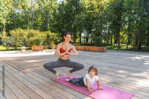 Beautiful young woman and charming little girl are smiling while doing yoga together. family relaxing and doing fitness at nature