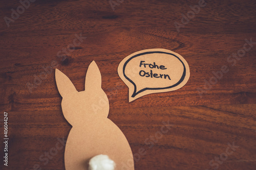 Umriss eines Osterhasen aus braunem Papier mit einer Sprechblase in der Frohe Ostern geschrieben steht auf einem Untergrund aus Holz. photo