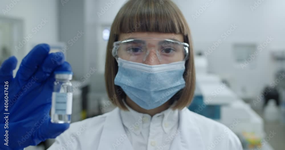 Portrait of female scientist in protective mask and googles looking to camera. Woman in white coat and latex gloves holding glass bottle with vaccine in laboratory. Zoom in. Portrait of female scientist in protective mask and googles looking to camera. Woman in white coat and latex gloves holding glass bottle with vaccine in laboratory. Zoom in.