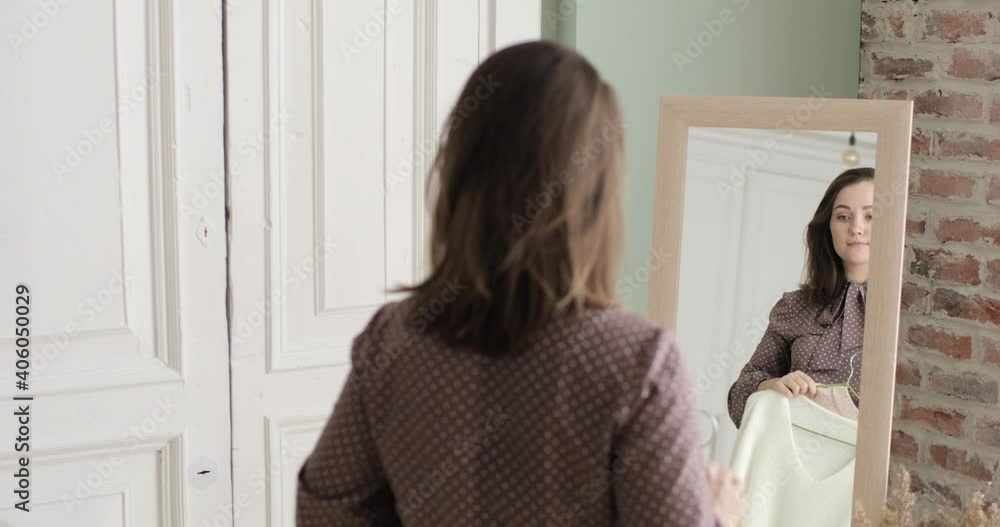Concept of everyday dilemma in outfit choosing. Young woman is trying two sweaters and looking at mirror, back view, shot in mirror reflection. The buyer chooses clothes in store, fashion boutique. Concept of everyday dilemma in outfit choosing. Young woman is trying two sweaters and looking at mirror, back view, shot in mirror reflection. The buyer chooses clothes in store, fashion boutique.