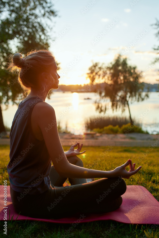 Fototapeta premium Side view of a young woman wearing sport clothes practicing yoga, sitting in lotus pose and meditating on the background of nature