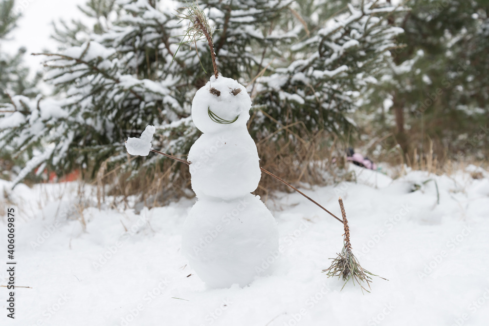 In the snow-covered forest stands a smiling snowman. Snowman shows like, in the other hand he has a spruce branch