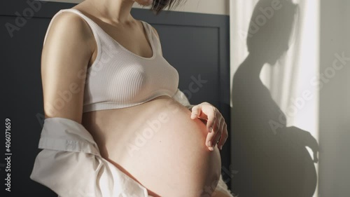 Young pregnant girl in white underwear and white shirt near a white wall in the sun