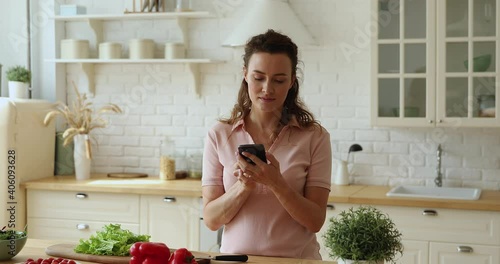 Woman standing in kitchen distracted from food preparation hold smartphone leaves voice message, search recipes on internet use speakerphone voice recognition virtual assistant, modern tech concept