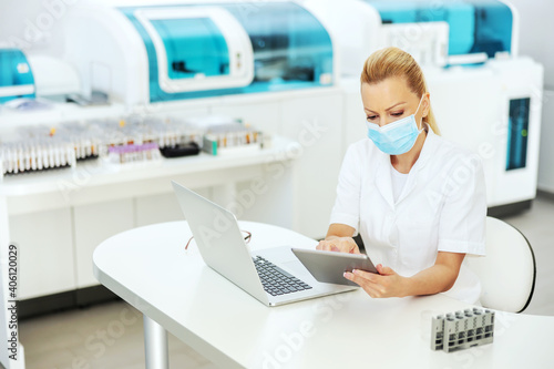 Attractive blond lab assistant in coat with sterile surgical mask on sitting in laboratory  entering test results on tablet. Research for cure for covid 19 concept.