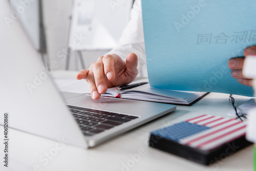 cropped view of interpreter working on laptop while holding paper with chinese hieroglyphs near digital translator on blurred foreground