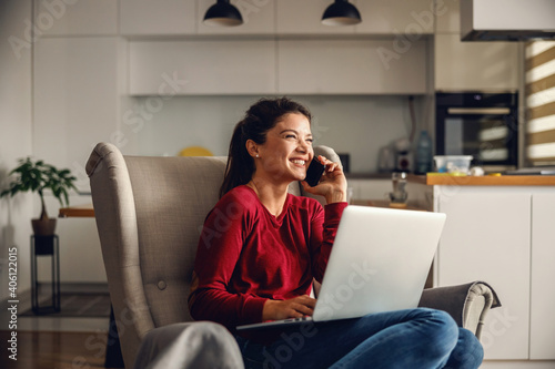 Smiling young woman sitting at home and having phone conversation with her boss. In lap she is holding laptop. Remote business during corona virus concept.