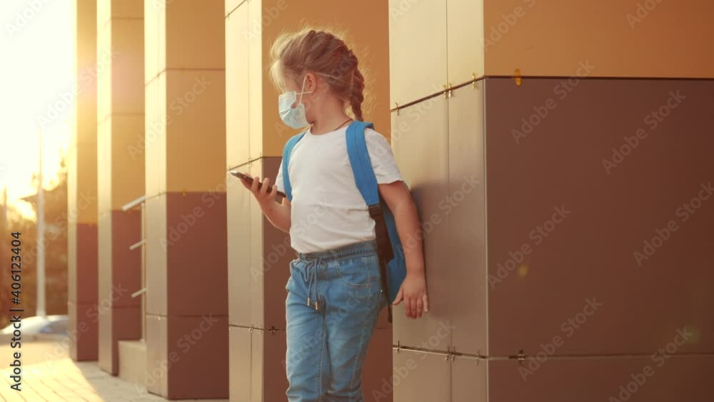 schoolgirl in a protective mask. child with a backpack stands near the school with a smartphone. schoolgirl in a protective mask outdoors. education coronavirus pandemic covid 19 concept schoolgirl in a protective mask. child with a backpack stands near the school with a smartphone. schoolgirl in a protective mask outdoors. education coronavirus pandemic covid 19 concept