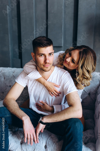 Studio photo session of a couple in love at the sofa. Incendiary meeting of a guy and a girl. Blonde in a white shirt. Grey background.