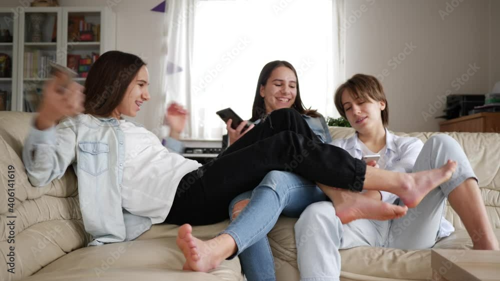 Teenager girls triplet sisters at home watch smart phones funny fighting with legs on couch Teenager girls triplet sisters at home watch smart phones funny fighting with legs on couch