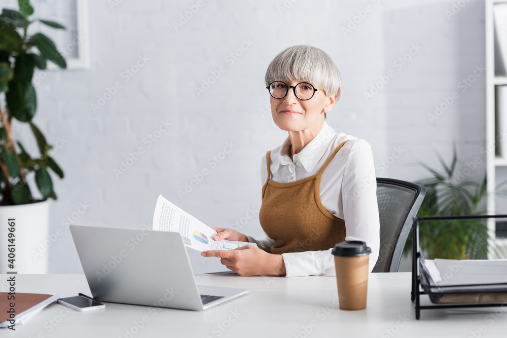 mature team leader sitting at desk and holding papers with charts and graphs in office mature team leader sitting at desk and holding papers with charts and graphs in office