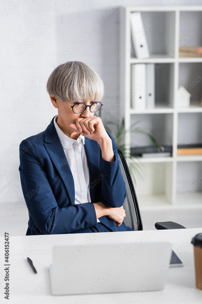 middle aged team leader in glasses covering mouth with clenched fist and looking at laptop middle aged team leader in glasses covering mouth with clenched fist and looking at laptop