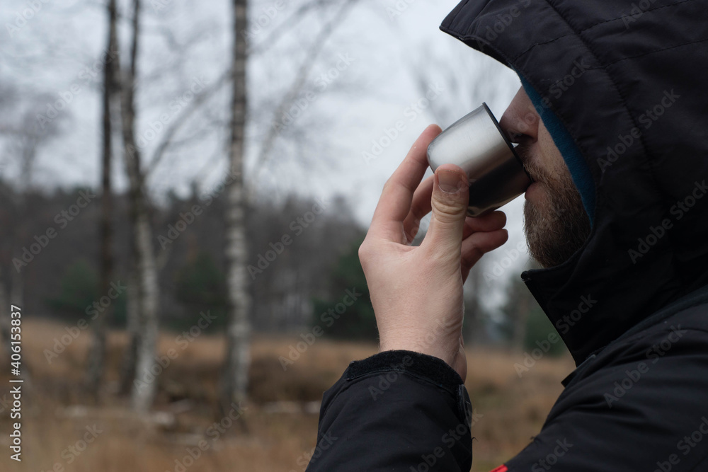 Young caucasian man, traveler in hooded winter jacket sipping hot tea straight from thermos cup in natural scenery. Survival and travel equipment useful during cold weather.