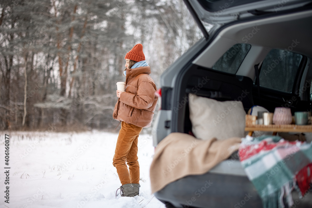 Woman with hot drink staying near car trunk in winter forest