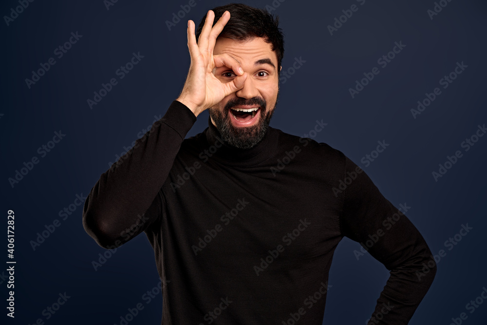 Cheeful, guy with beard wearingn black high neck sweater, showing okay, excellent, good gesture, make circle over eye and look through, with happy smile, agree, give recommendation, approve choice