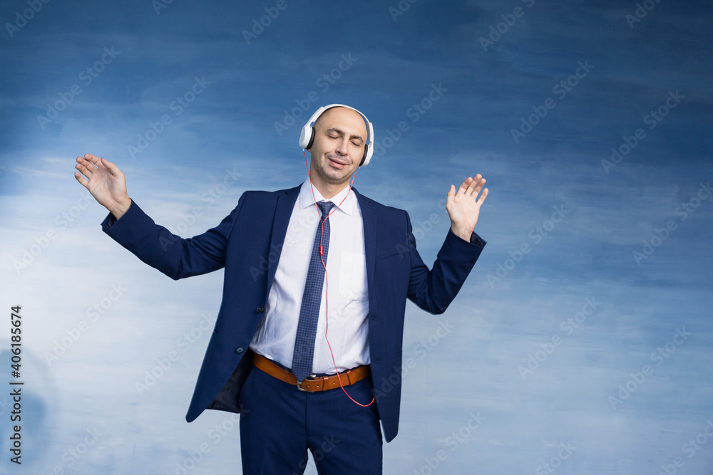 A bald man in a jacket and tie with eyes closed dances and enjoys music in large headphones on a blue background A bald man in a jacket and tie with eyes closed dances and enjoys music in large headphones on a blue background