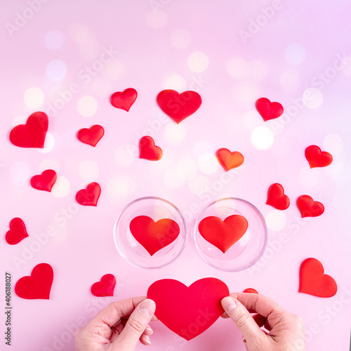 Hands hold a red heart shape, two glasses with hearts and white and red heart shapes on a pink background with bokeh, top view, copy space, square frame. Valentine's Day concept