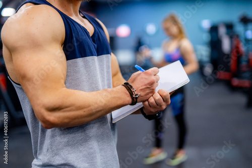 Selective focus on muscular hands with clipboard. Unrecognizible fitness trainer gives recommendations for better results. Client girl on blurred background.