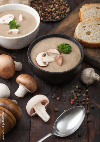 Ceramic bowl plates of creamy chestnut champignon mushroom soup with spoon, pepper and kitchen cloth on dark wooden background.
