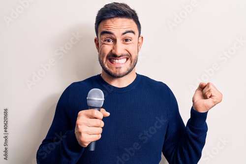 Young handsome singer man with beard singing song using microphone over white background screaming proud, celebrating victory and success very excited with raised arm