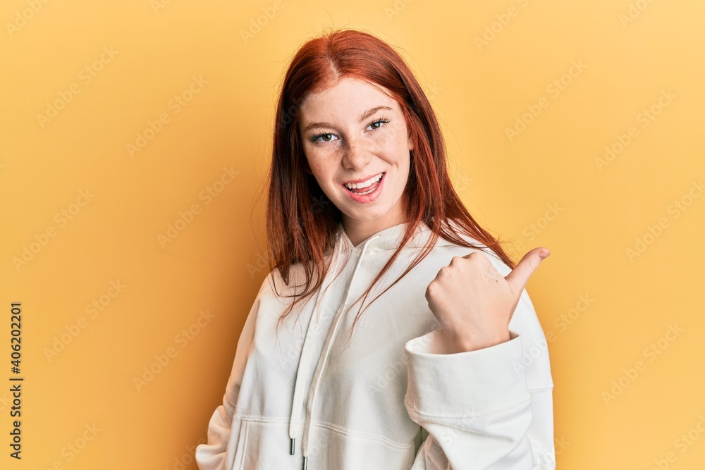 Young red head girl wearing casual sweatshirt smiling with happy face looking and pointing to the side with thumb up. Young red head girl wearing casual sweatshirt smiling with happy face looking and pointing to the side with thumb up.