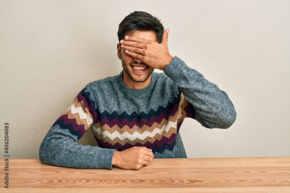 Young handsome man wearing casual sweater sitting on the table smiling and laughing with hand on face covering eyes for surprise. blind concept. Young handsome man wearing casual sweater sitting on the table smiling and laughing with hand on face covering eyes for surprise. blind concept.