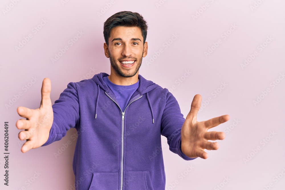 Young handsome man wearing casual sweatshirt looking at the camera smiling with open arms for hug. cheerful expression embracing happiness. Young handsome man wearing casual sweatshirt looking at the camera smiling with open arms for hug. cheerful expression embracing happiness.