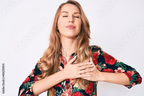 Young caucasian woman with blond hair wearing elegant floral shirt smiling with hands on chest, eyes closed with grateful gesture on face. health concept.