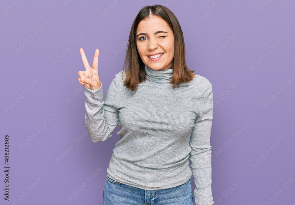 Young beautiful woman wearing casual turtleneck sweater smiling with happy face winking at the camera doing victory sign with fingers. number two. Young beautiful woman wearing casual turtleneck sweater smiling with happy face winking at the camera doing victory sign with fingers. number two.