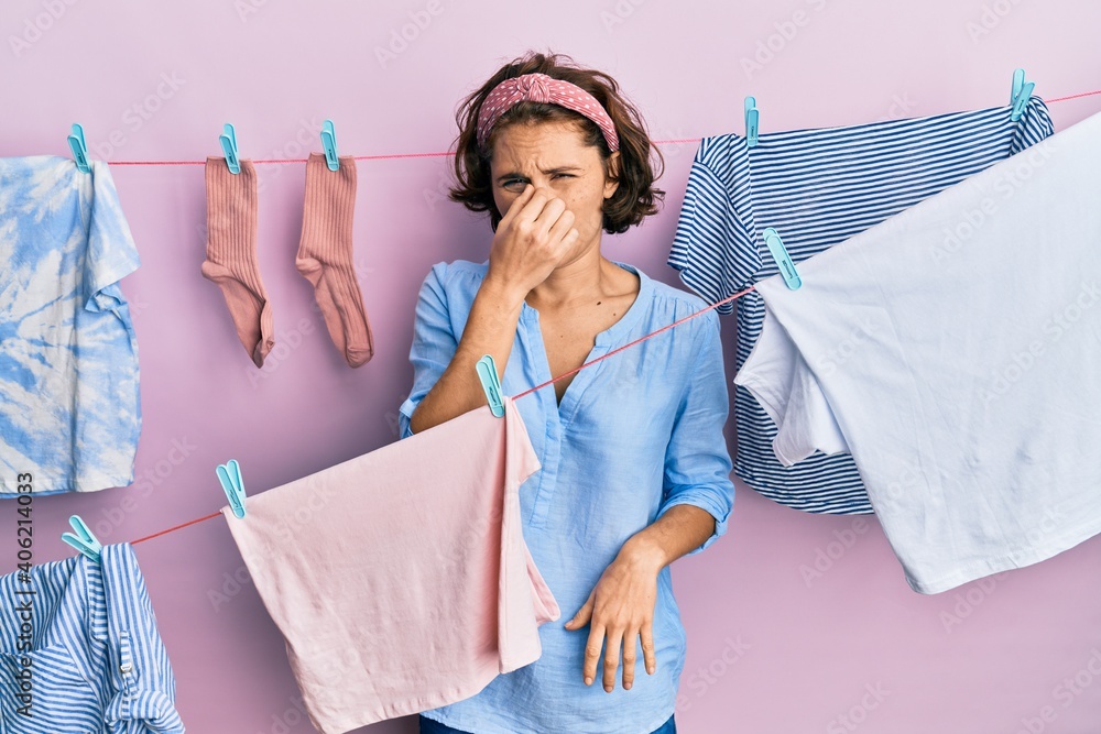 Young brunette woman doing laundry around string hangs smelling something stinky and disgusting, intolerable smell, holding breath with fingers on nose. bad smell - obrazy, fototapety, plakaty 