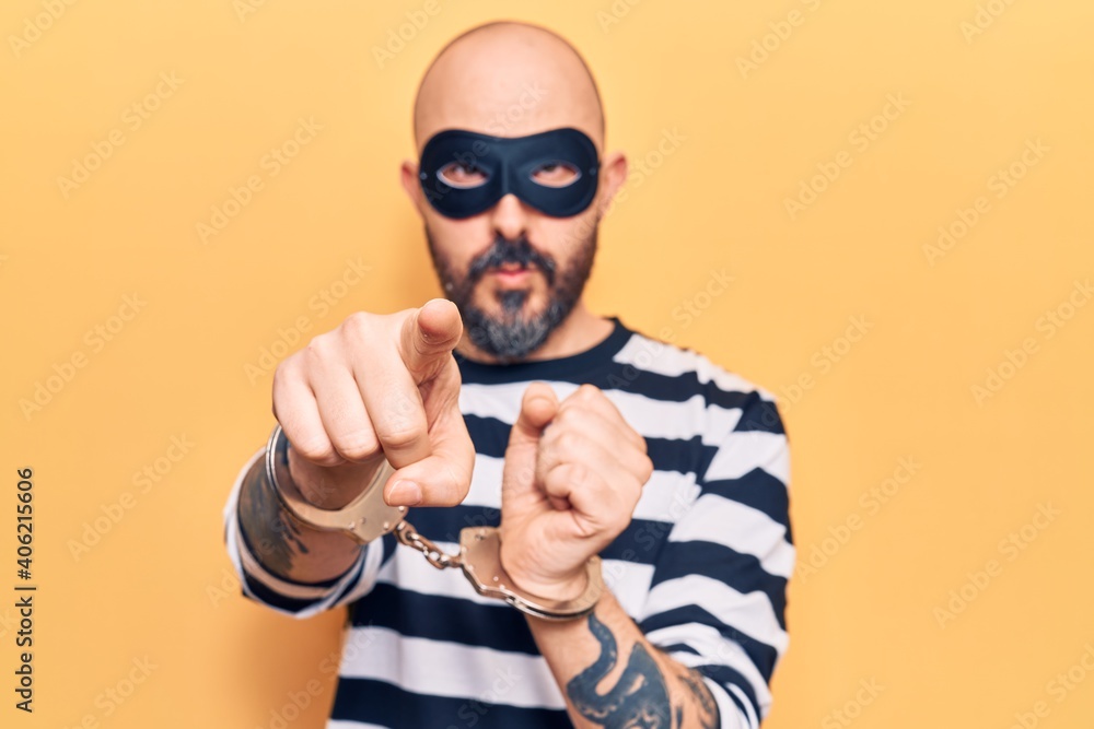 Young handsome man wearing burglar mask and handcuffs pointing with finger to the camera and to you, confident gesture looking serious Young handsome man wearing burglar mask and handcuffs pointing with finger to the camera and to you, confident gesture looking serious