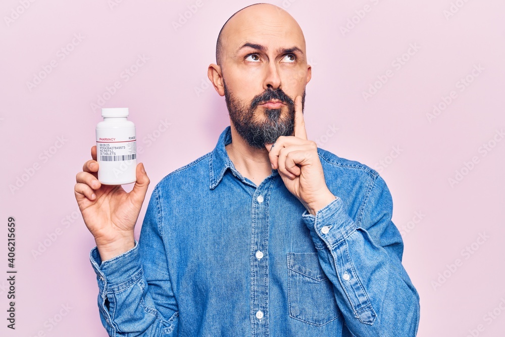 Young handsome man holding pills serious face thinking about question with hand on chin, thoughtful about confusing idea Young handsome man holding pills serious face thinking about question with hand on chin, thoughtful about confusing idea