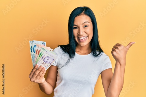 Beautiful hispanic woman holding singapore dollars banknotes pointing thumb up to the side smiling happy with open mouth
