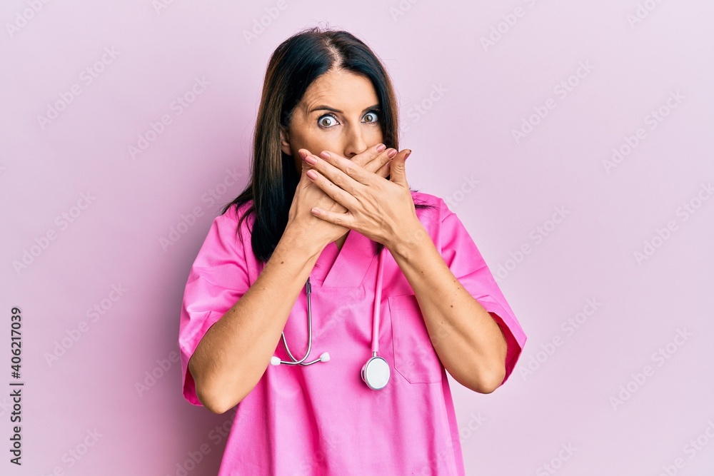 Middle age brunette woman wearing doctor uniform and stethoscope shocked covering mouth with hands for mistake. secret concept. Middle age brunette woman wearing doctor uniform and stethoscope shocked covering mouth with hands for mistake. secret concept.
