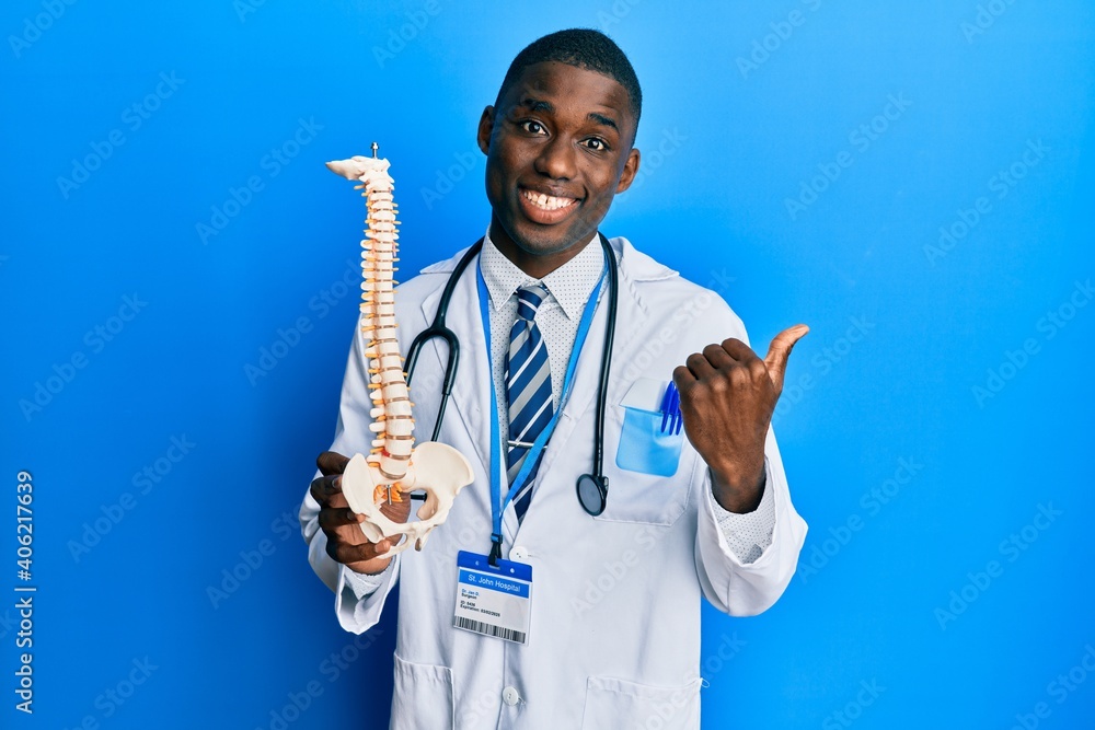 Young african american doctor man holding anatomical model of spinal column pointing thumb up to the side smiling happy with open mouth Young african american doctor man holding anatomical model of spinal column pointing thumb up to the side smiling happy with open mouth