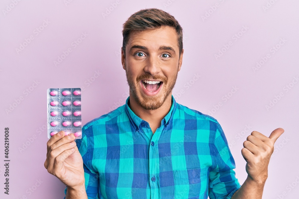 Handsome caucasian man holding pills pointing thumb up to the side smiling happy with open mouth Handsome caucasian man holding pills pointing thumb up to the side smiling happy with open mouth