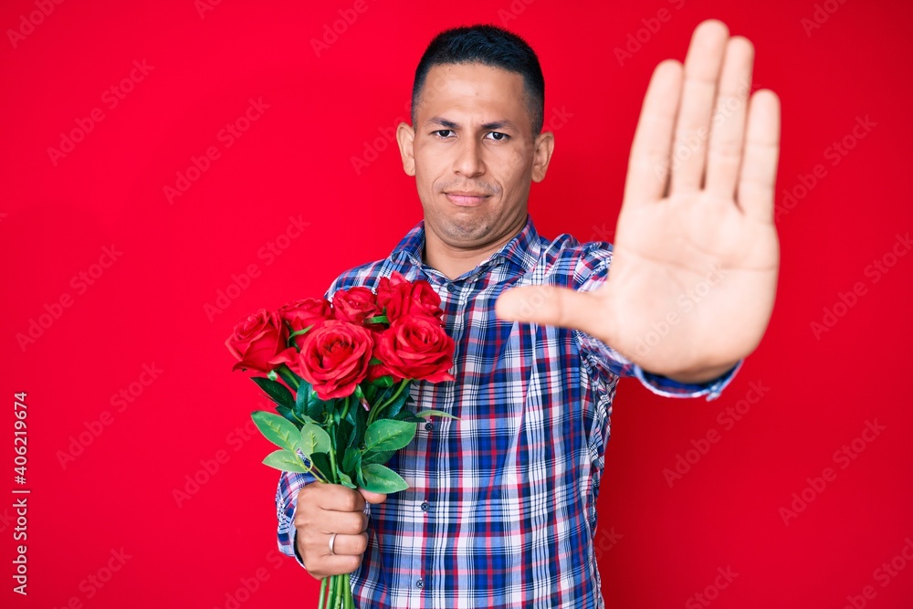 Young handsome latin man holding flowers with open hand doing stop sign with serious and confident expression, defense gesture Young handsome latin man holding flowers with open hand doing stop sign with serious and confident expression, defense gesture