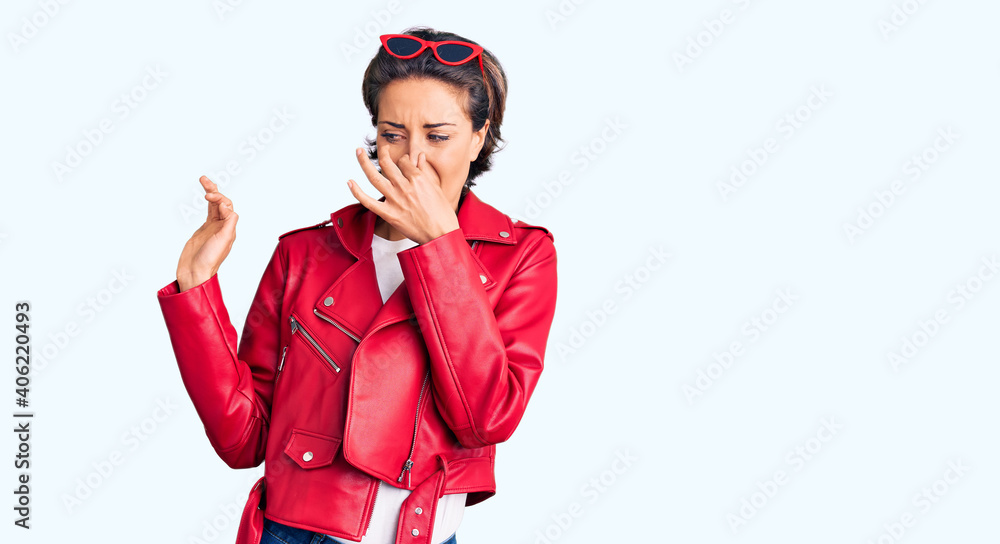 Young beautiful woman wearing red leather jacket smelling something stinky and disgusting, intolerable smell, holding breath with fingers on nose. bad smell Young beautiful woman wearing red leather jacket smelling something stinky and disgusting, intolerable smell, holding breath with fingers on nose. bad smell