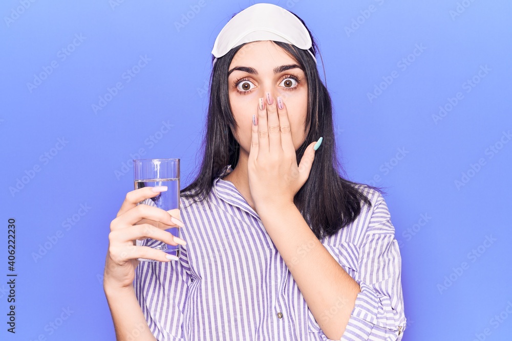 Young beautiful girl wearing sleep mask drinking glass of water covering mouth with hand, shocked and afraid for mistake. surprised expression Young beautiful girl wearing sleep mask drinking glass of water covering mouth with hand, shocked and afraid for mistake. surprised expression