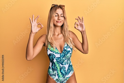 Beautiful blonde young woman wearing swimsuit and sunglasses relax and smiling with eyes closed doing meditation gesture with fingers. yoga concept.