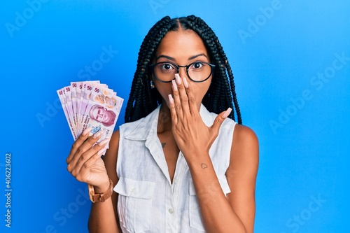 Beautiful hispanic woman holding 50 mexican pesos banknotes covering mouth with hand, shocked and afraid for mistake. surprised expression