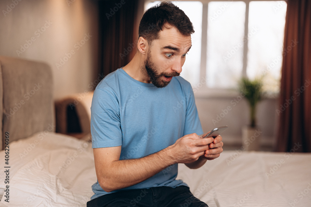 Young man with beard surprised shocked whilst received sending message on phone while sitting in bed. Young man with beard surprised shocked whilst received sending message on phone while sitting in bed.
