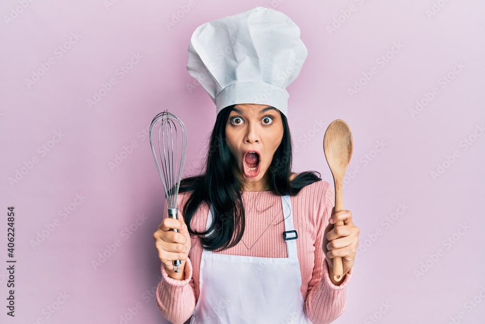 Fototapeta premium Young hispanic woman wearing baker uniform holding spoon and whisk afraid and shocked with surprise and amazed expression, fear and excited face.
