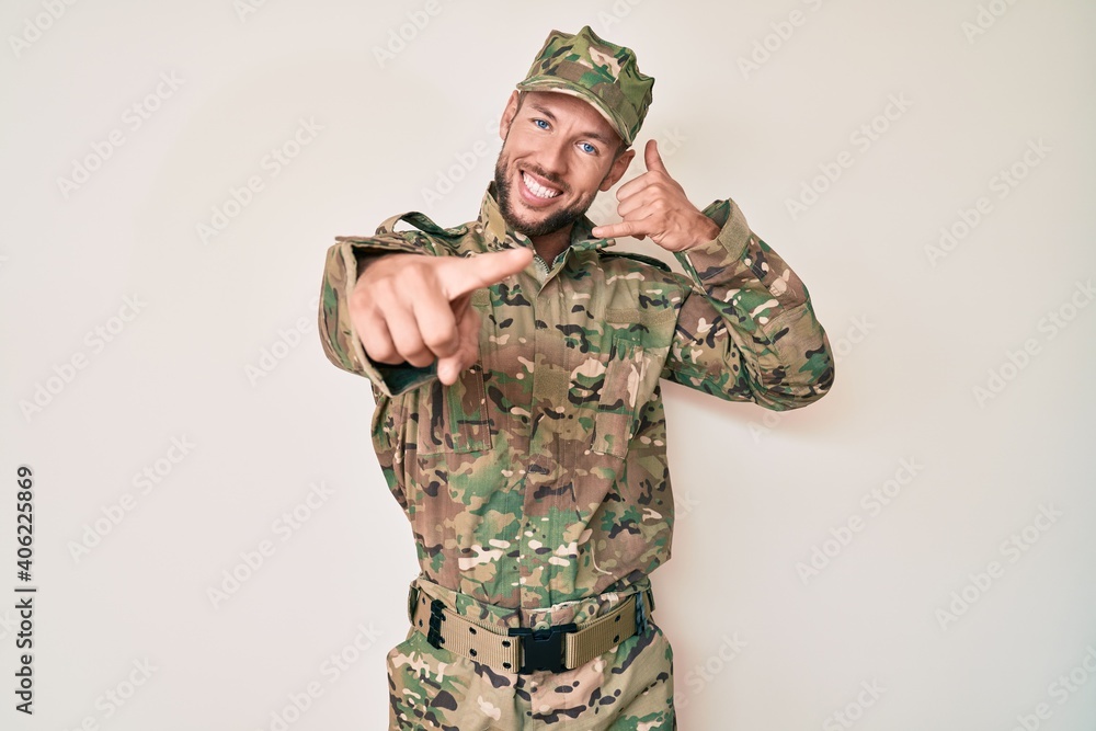 Young caucasian man wearing camouflage army uniform smiling doing talking on the telephone gesture and pointing to you. call me. Young caucasian man wearing camouflage army uniform smiling doing talking on the telephone gesture and pointing to you. call me.