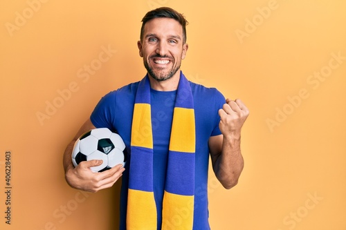 Handsome man with beard football hooligan cheering game holding ball screaming proud, celebrating victory and success very excited with raised arm