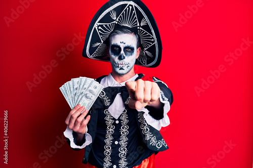 Young man wearing mexican day of the dead costume holding dollars pointing with finger to the camera and to you, confident gesture looking serious