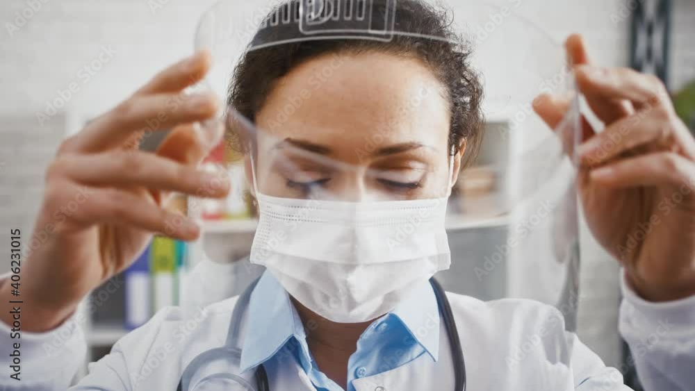 Doctor woman in protective mask is putting on a face shield and looking at you while standing in cabinet of modern clinic Doctor woman in protective mask is putting on a face shield and looking at you while standing in cabinet of modern clinic